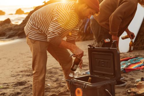 Two adult men on a sandy beach with an open cooler filled with bottled beer, next to a surfboard and rocks during the day.