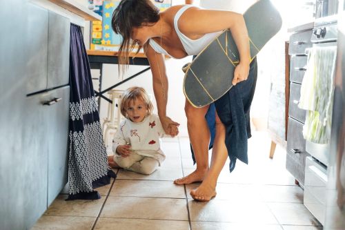 A mother in a tank top holds a skateboard while holding hands with her toddler in a sunlit kitchen. The scene conveys a relaxed, family moment.