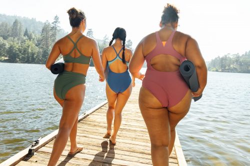 Three women in swimsuits walk on a wooden dock toward a lake with trees in the background, each holding a yoga mat.