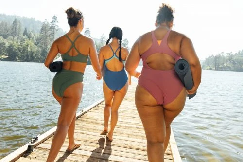 Three women in swimsuits walk on a wooden dock toward a lake with trees in the background, each holding a yoga mat.