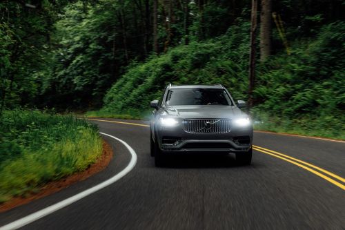 Gray SUV driving on an asphalt road surrounded by lush green trees and foliage. Car headlights are on. Road markings visible.