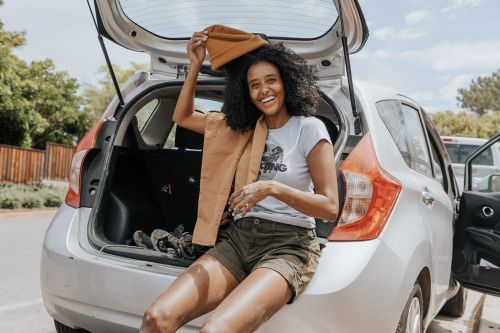 Smiling woman sits in the open trunk of a silver car, holding a brown hat and vest, with trees and a white car visible in the background.