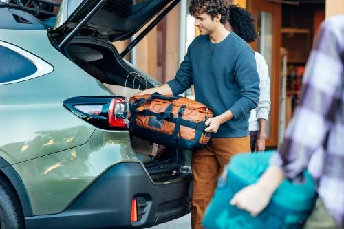 Olive-green SUV with its trunk open, a man loading a brown duffel bag, and two other people standing nearby, all facing the vehicle.