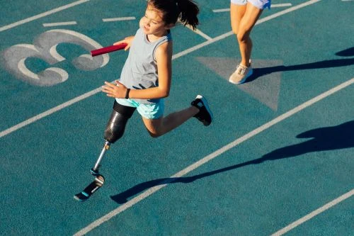 A young girl with a prosthetic leg is running on a blue track with a relay baton in hand, a second person is running behind her.