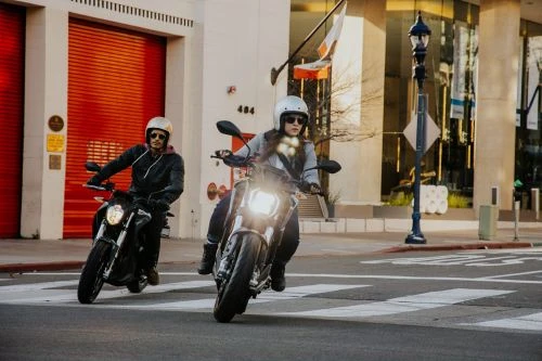 Two motorcycle riders wearing helmets driving on a city street with a building with orange doors in the background on a sunny day.