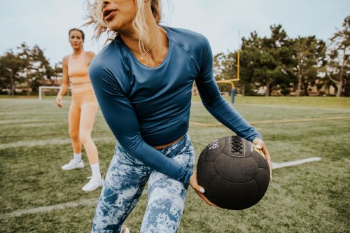 Two women work out on a sports field, one holding a medicine ball in the foreground and one in the background.