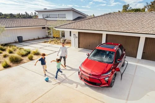Red electric SUV charges in driveway as a father watches two children playing soccer on a sunny day in front of a modern house.