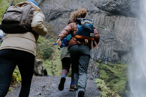 Three people with backpacks walk away on a path in a natural environment. A waterfall and rock wall are in the background.