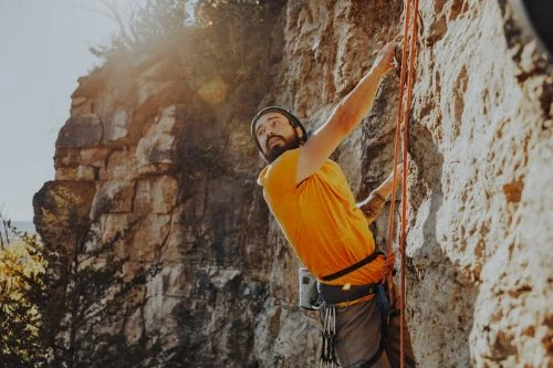 Bearded adult rock climber in protective gear ascending a textured rock face with a rope in bright sunlight, outdoor setting.