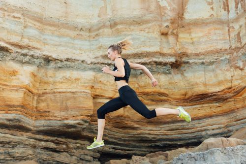Athletic woman leaps against layered rock formation. Wearing black athletic clothing and lime green shoes. Dynamic fitness and outdoor training.