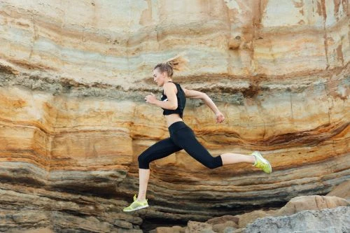 Athletic woman leaps against layered rock formation. Wearing black athletic clothing and lime green shoes. Dynamic fitness and outdoor training.