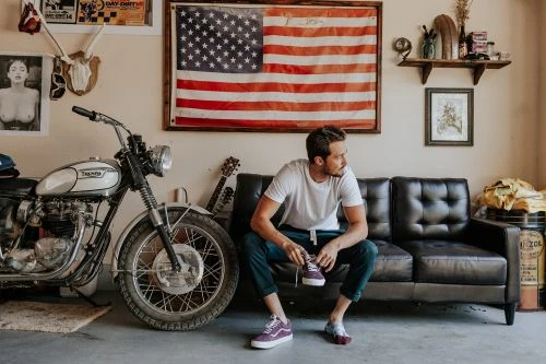 Man sits on a black leather sofa indoors near a motorcycle and American flag, wearing casual clothes and holding a purple sneaker.
