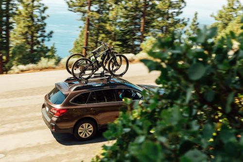 Brown SUV with two mountain bikes on the roof rack travels along a road with green foliage and ocean in the background.