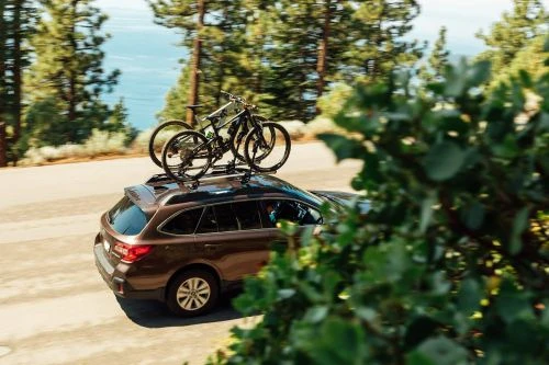 Brown SUV with two mountain bikes on the roof rack travels along a road with green foliage and ocean in the background.
