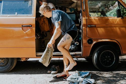 Man stands near an open vintage van, barefoot with visible tattoos, holding clothes, with trees visible in the background on a sunny day.
