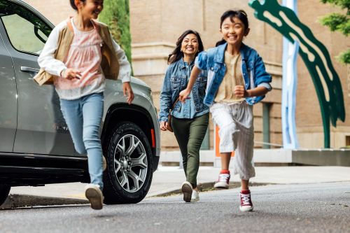 Two young girls and an adult woman running towards the camera on a city street next to a gray SUV with a building backdrop.