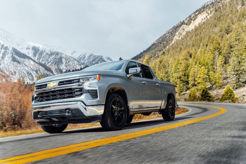 Silver Chevrolet Silverado pickup truck drives on a paved mountain road with snow-capped peaks in the background, showcasing automotive engineering.