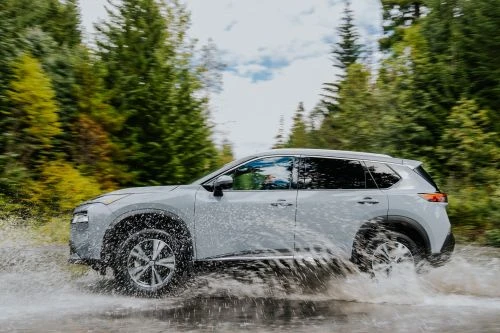 Silver SUV driving through water on a dirt road, with green trees and a cloudy sky in the background, showcasing automotive capabilities.