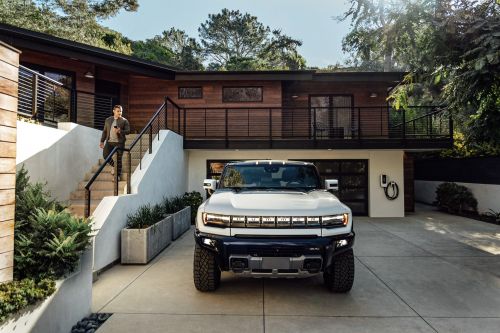 White Hummer EV parked on a concrete driveway in front of a modern two-story house with wood siding. A man descends the stairs.