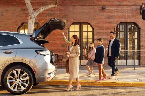 Woman in beige coat reaching toward an open SUV trunk while family walks past brick building
