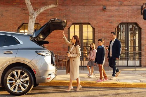 Woman in beige coat reaching toward an open SUV trunk while family walks past brick building