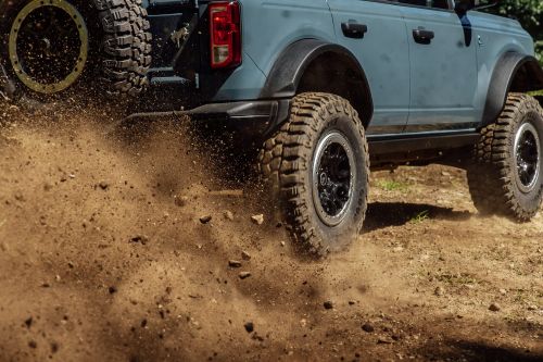 Off-road truck in action, blue Ford Bronco driving on a dirt road kicking up dirt and dust with rugged terrain in the background.