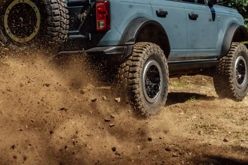 Off-road truck in action, blue Ford Bronco driving on a dirt road kicking up dirt and dust with rugged terrain in the background.