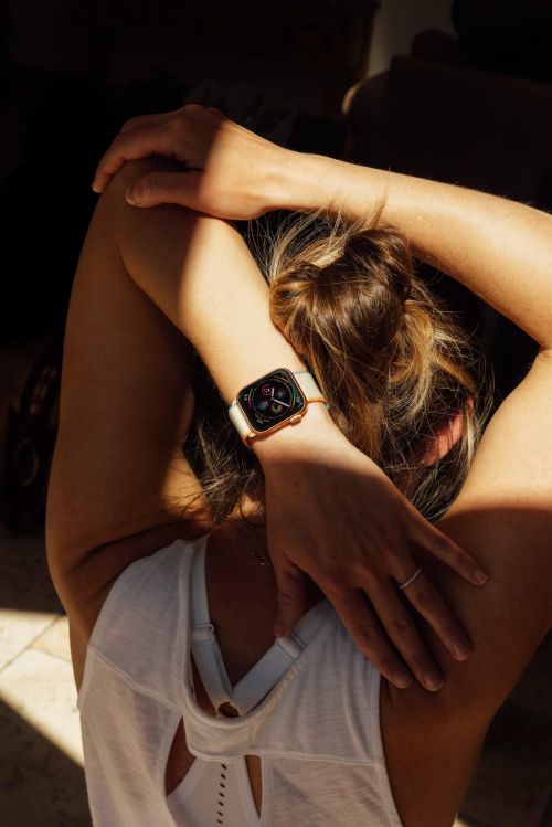Woman stretches arms behind back, wearing a white tank top and a smartwatch with a light-colored band in indoor lighting.