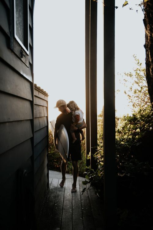 Adult carries child with surfboard on path near house and trees. Man holds young kid on hip in bright daylight.