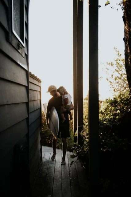 Adult carries child with surfboard on path near house and trees. Man holds young kid on hip in bright daylight.