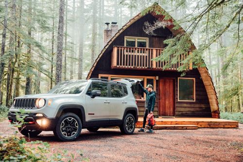 Silver Jeep SUV parked near an A-frame cabin with a man standing by the open rear hatch in a forest environment.