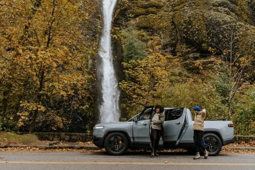 Two adults pose in front of a gray Rivian pickup truck at the base of a forest waterfall, with a road in the foreground.