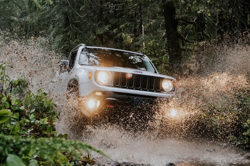 White Jeep driving through a puddle in a forest, water splashes everywhere, headlights on, off-road vehicle in nature.