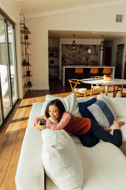 Woman relaxes on white sofa in sunlit living room with open kitchen, wood floors, decorative pillows, indoor plants and sliding glass doors.