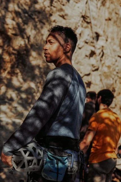 Man in climbing gear stands before a rock wall; wearing a grey shirt and blue climbing bag; light and shadow dapple the scene.