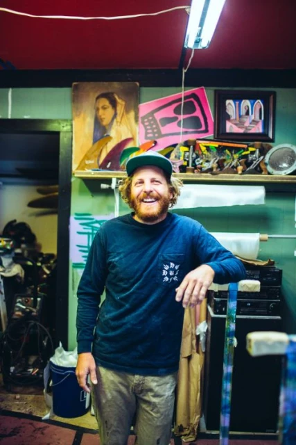 Portrait of a smiling man with a beard in front of paintings and shelves in an art studio with indoor lighting.