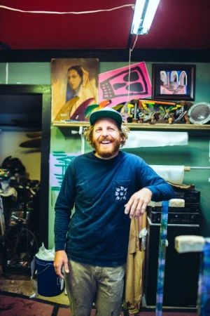 Portrait of a smiling man with a beard in front of paintings and shelves in an art studio with indoor lighting.