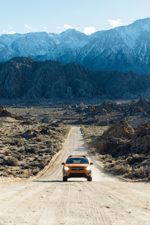 Orange SUV driving along a dirt road through a desert landscape, with snow-capped mountains in the background under a blue sky.