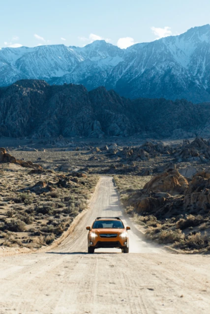 Orange SUV driving along a dirt road through a desert landscape, with snow-capped mountains in the background under a blue sky.