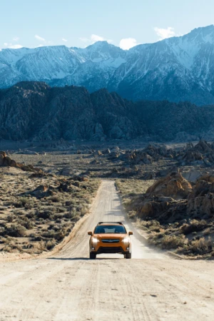 Orange SUV driving along a dirt road through a desert landscape, with snow-capped mountains in the background under a blue sky.