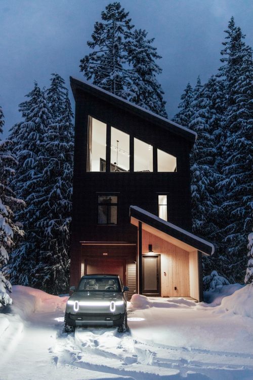Modern architectural home with angled roof and front-facing electric SUV parked in snow next to trees at dusk.