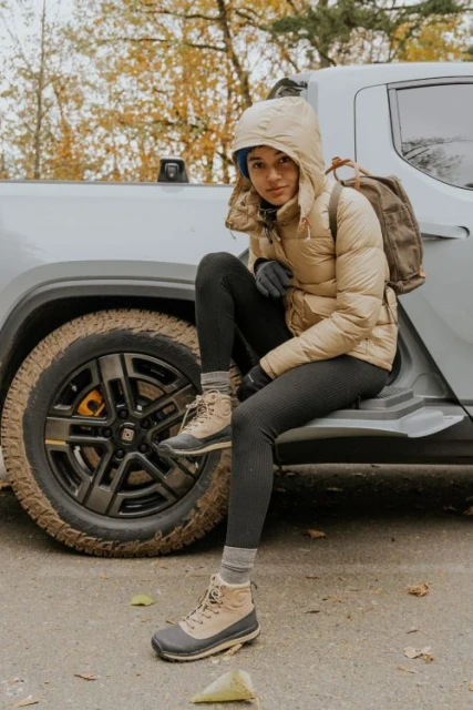 A woman in a puffer jacket sits on the bumper of a Rivian truck near a forest, wearing hiking boots and a backpack. 