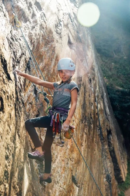 A young girl in a helmet is rock climbing on a tan rock wall with climbing equipment and ropes in an outdoor setting with greenery.
