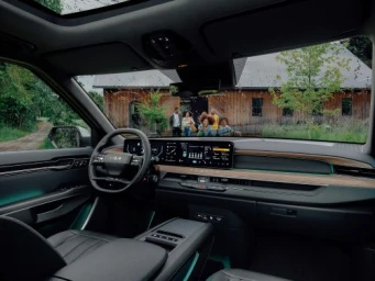 Interior view of a modern car dashboard with a wide digital display, steering wheel, and a group of people visible through the windshield.