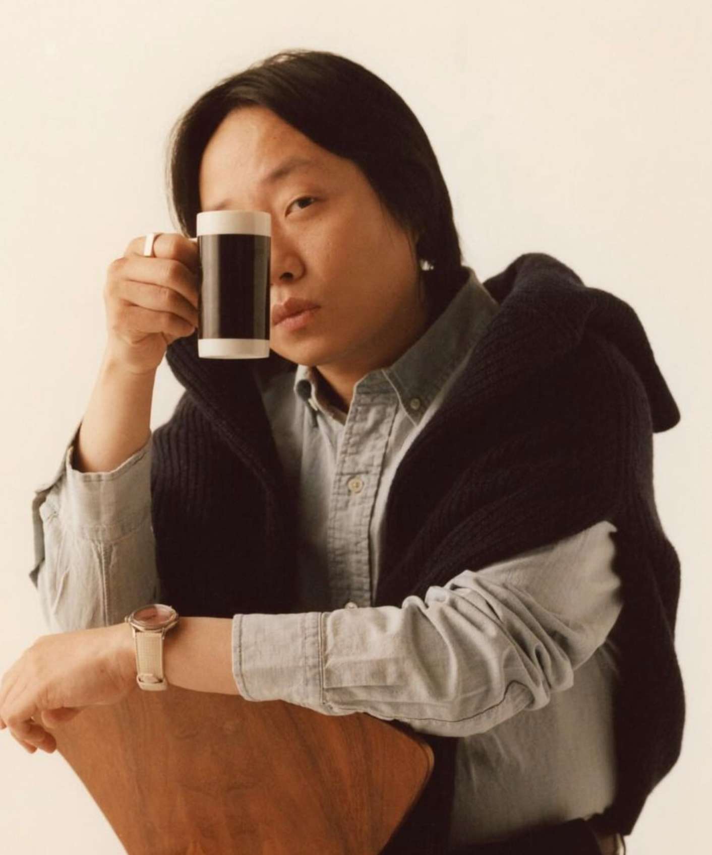 Portrait of an Asian adult in a blue shirt, wearing a wristwatch, holding a black and white mug against a neutral background.