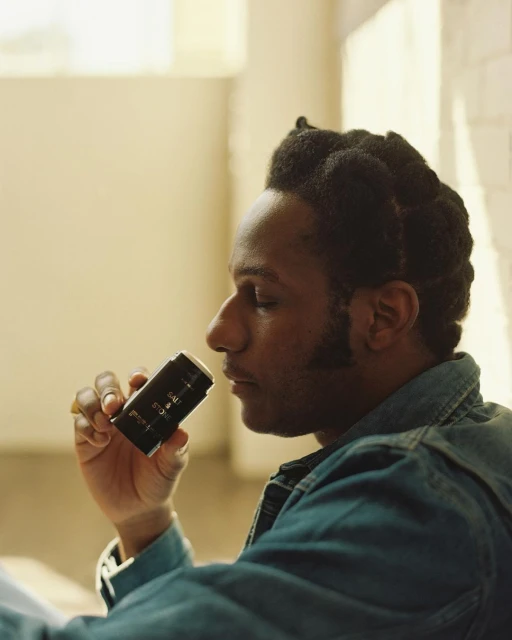 A Black male with dark hair and a blue denim jacket is smelling a black and gold deodorant stick indoors, near a window.