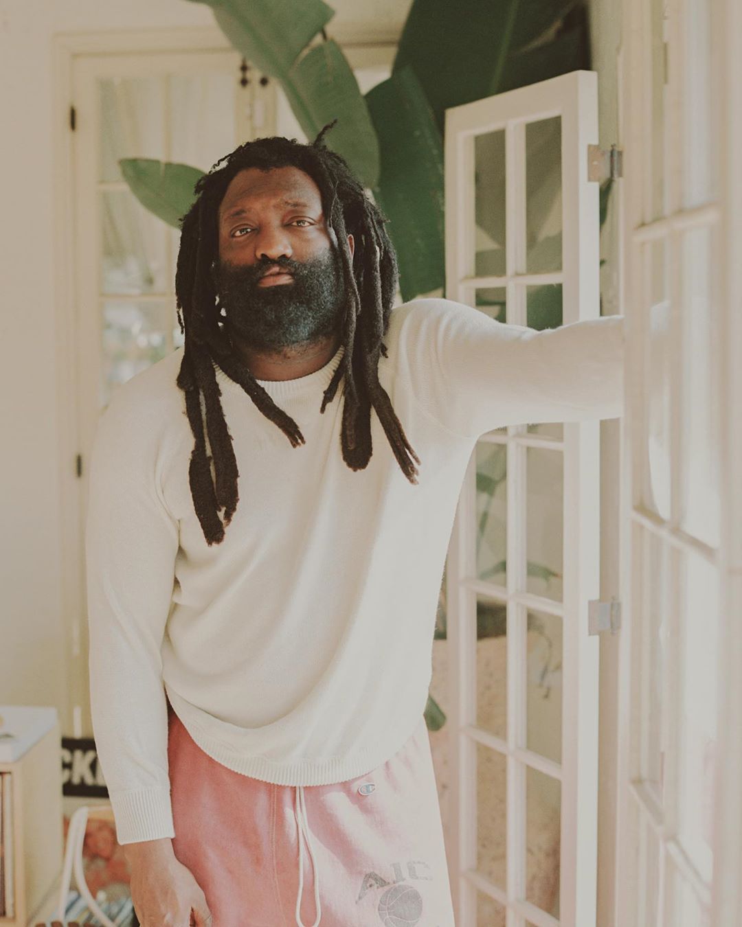 Portrait of an adult man with dreadlocks wearing a white sweater and pink sweatpants, posing indoors in front of white doors and plants.