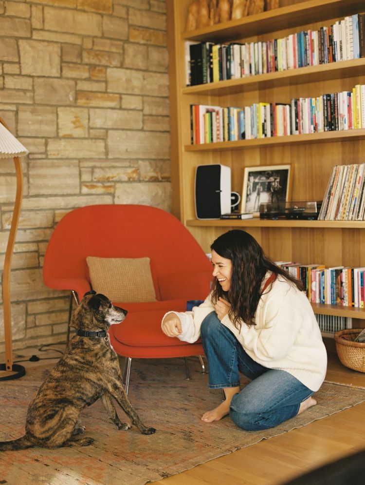 A young woman with long hair interacts with a brindle dog indoors near a red chair, stone wall, and wooden bookshelf filled with books.