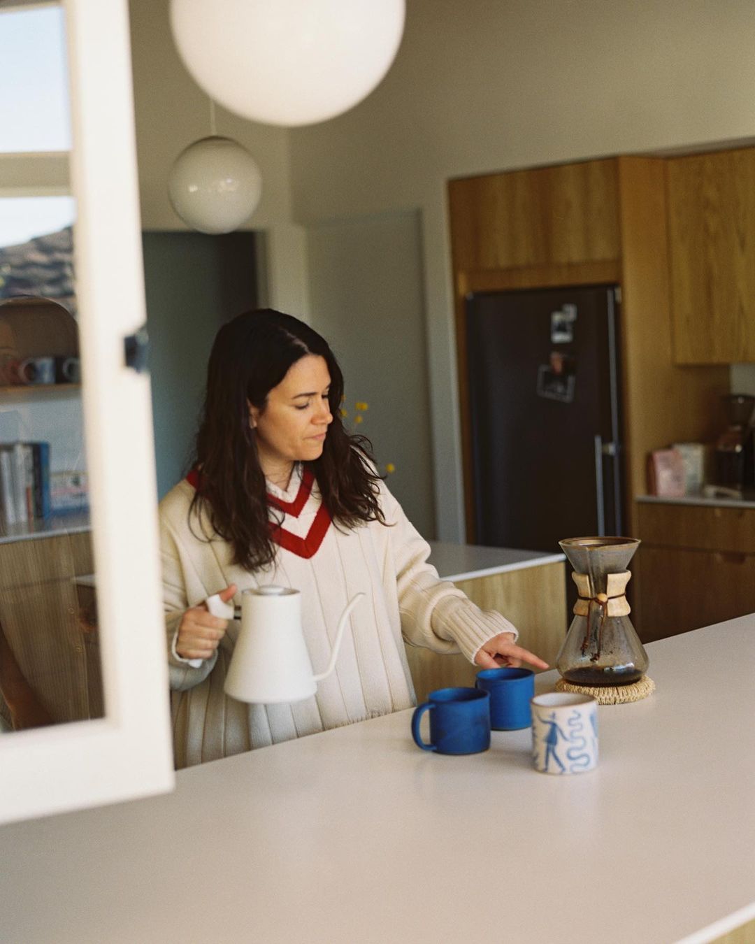 Woman preparing pour-over coffee at a white kitchen counter with mugs, a kettle, and kitchen cabinets visible in soft, natural light.