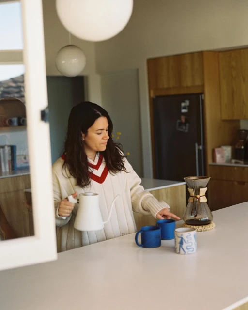 Woman preparing pour-over coffee at a white kitchen counter with mugs, a kettle, and kitchen cabinets visible in soft, natural light.
